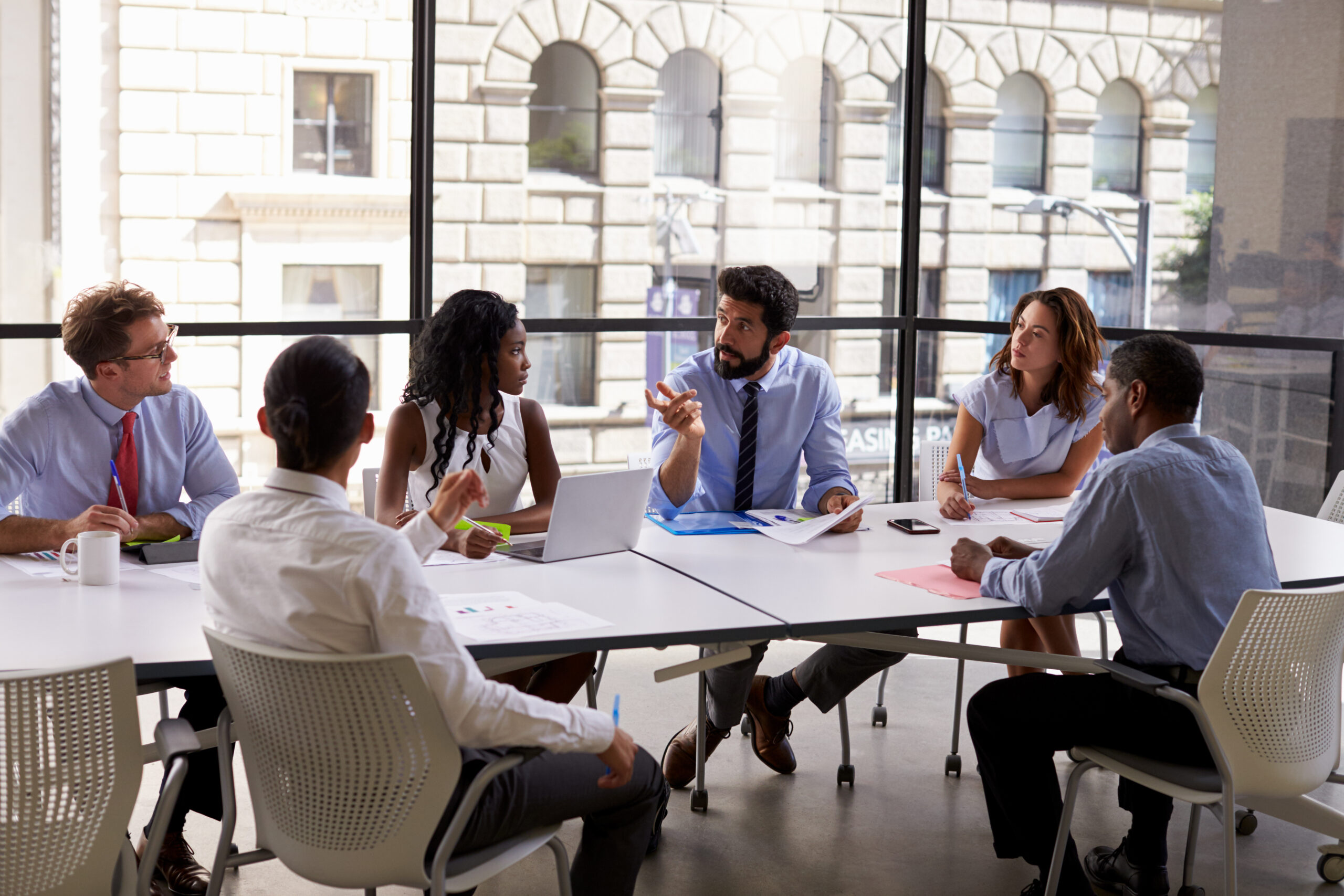 A diverse group of professionals engage in a meeting around a conference table. The setting is a modern office with large windows and urban views, conveying collaboration.